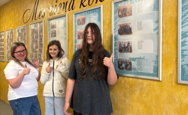 Skolniece ar skolotāju smaidot rāda īkšķus uz augšu gaišā, dzeltenā koridorī. Uz sienas liels uzraksts: 