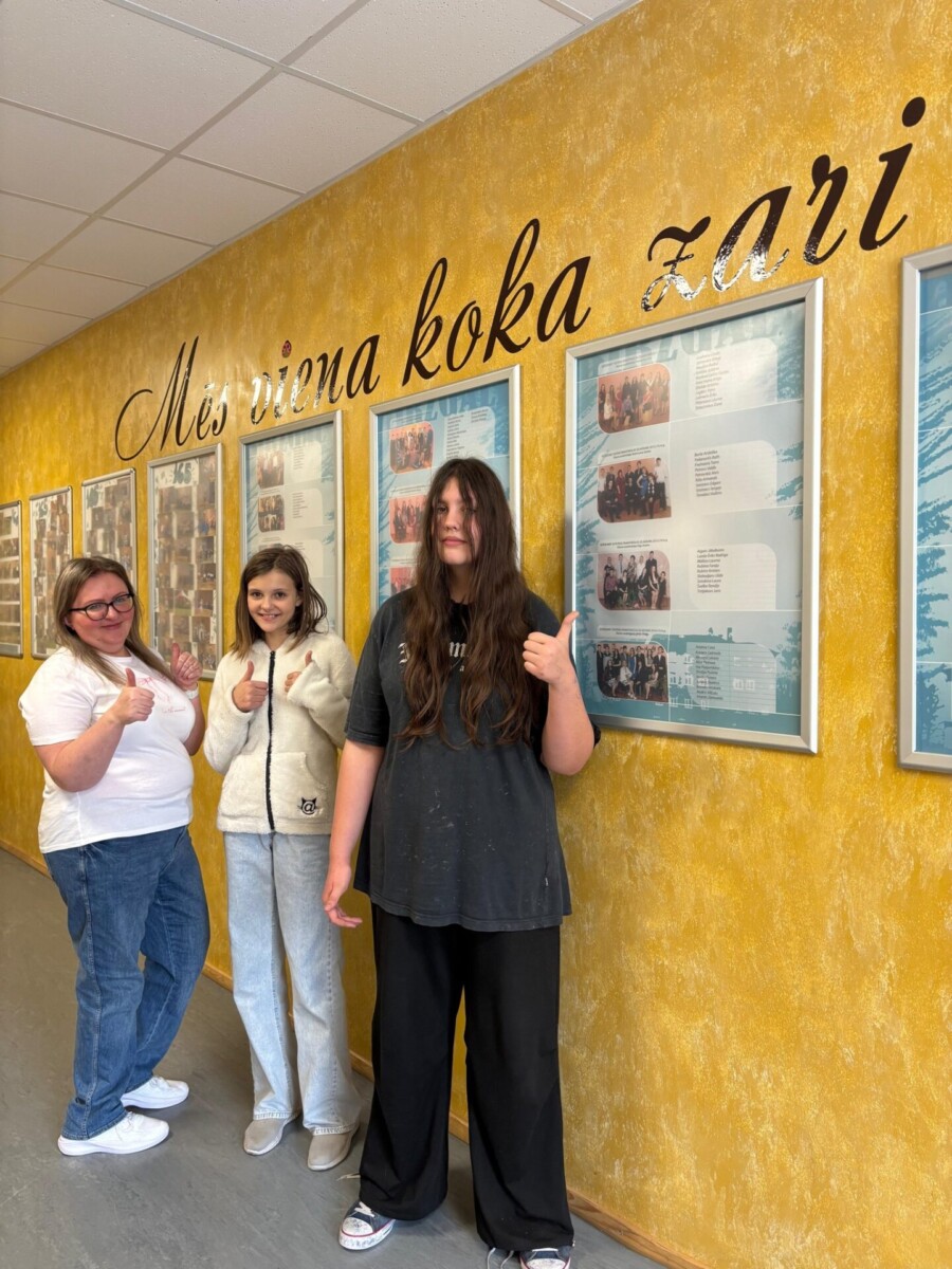 Skolniece ar skolotāju smaidot rāda īkšķus uz augšu gaišā, dzeltenā koridorī. Uz sienas liels uzraksts: 