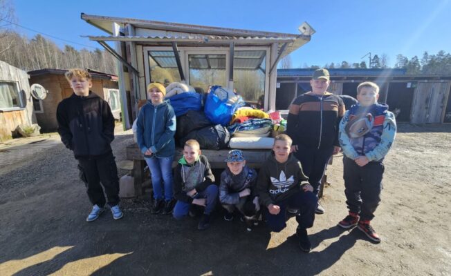 Astoņi bērni, septiņi zēni un viena meitene, pozē āra vidē. Viņiem blakus uz soliņa ir sakrautas drēbes un segas, bet fonā redzamas koka suņu būdas zem skaidrām zilām debesīm. 4.klases skolēni devušies uz dzīvnieku patversmi 