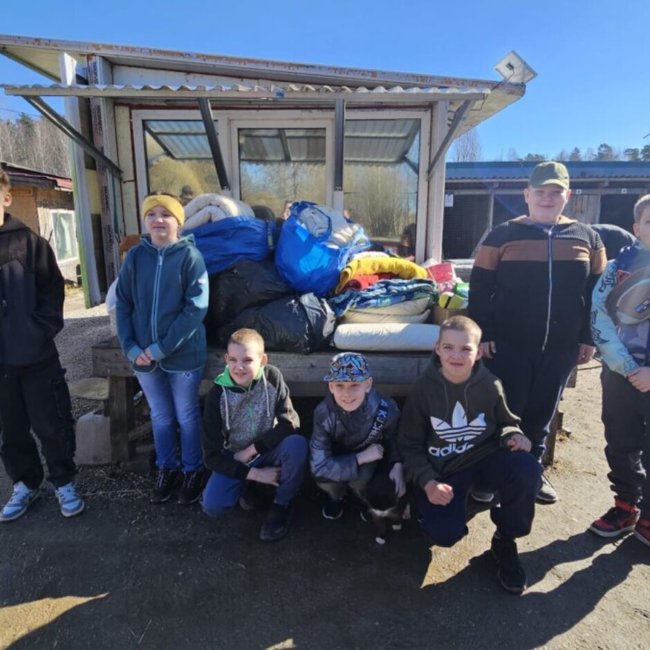 Astoņi bērni, septiņi zēni un viena meitene, pozē āra vidē. Viņiem blakus uz soliņa ir sakrautas drēbes un segas, bet fonā redzamas koka suņu būdas zem skaidrām zilām debesīm. 4.klases skolēni devušies uz dzīvnieku patversmi 