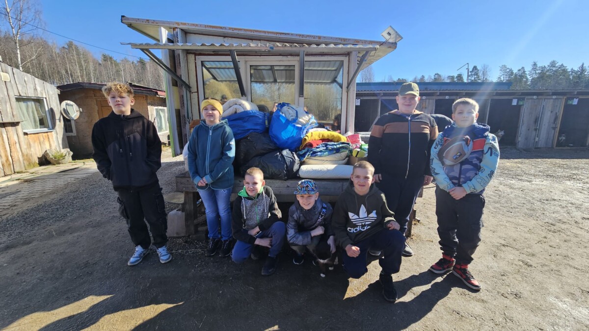 Astoņi bērni, septiņi zēni un viena meitene, pozē āra vidē. Viņiem blakus uz soliņa ir sakrautas drēbes un segas, bet fonā redzamas koka suņu būdas zem skaidrām zilām debesīm. 4.klases skolēni devušies uz dzīvnieku patversmi 