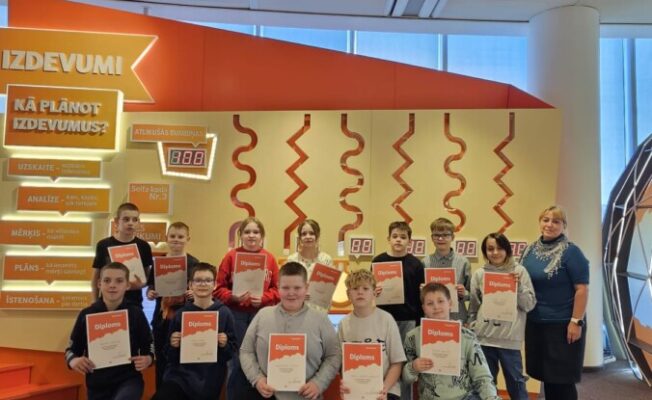 Divpadsmit skolēni un viena pieauguša sieviete smaida un pozē, turot 