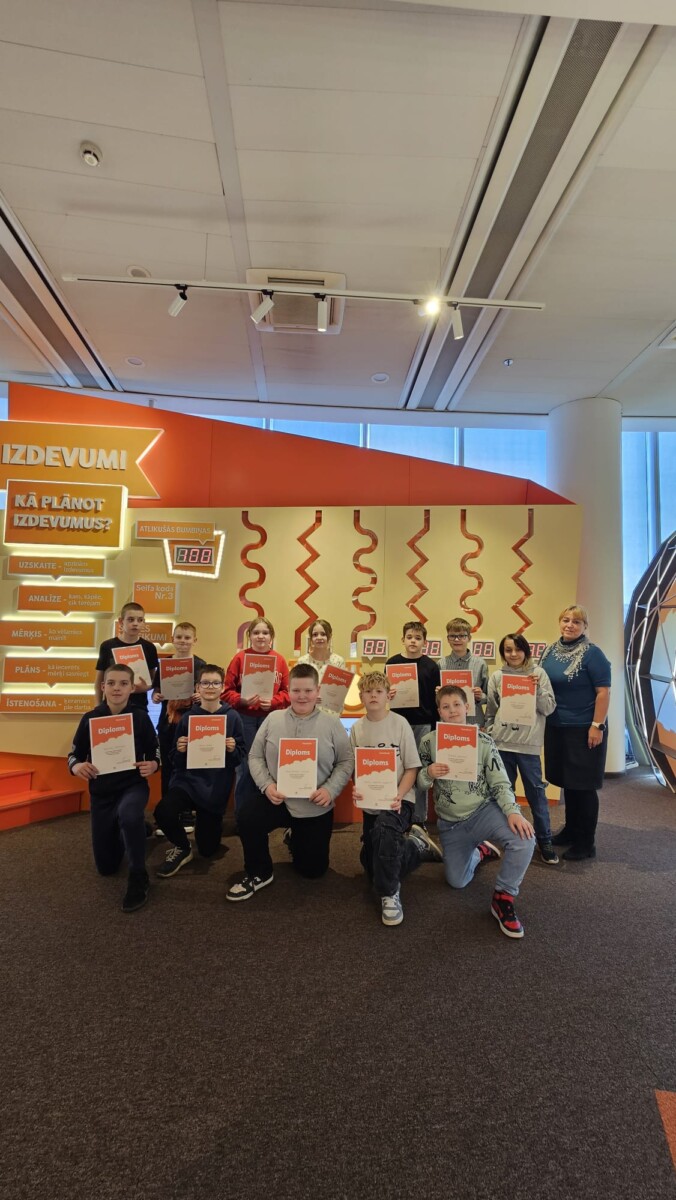 Divpadsmit skolēni un viena pieauguša sieviete smaida un pozē, turot 