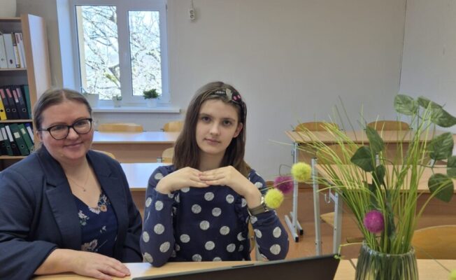 Sieviete un meitene sēž pie koka galda gaišā telpā. Sieviete ar brillēm un tumšu žaketi smaidot skatās kamerā. Meitene, uz galda nolikusi klēpjdatoru, salikusi rokas priekšā un skatās tieši kamerā. Uz galda stāv arī vāze ar zaļiem augiem un krāsainiem pomponu rotājumiem. Fonā redzams logs ar koku zariem un grāmatu plaukts. Meitene ir 4.klases skolniece Sofija Piļipenko, kura piedalās angļu valodas valsts olimpiādē, bet dāma ir viņas skolotāja Linda Liepiņa.