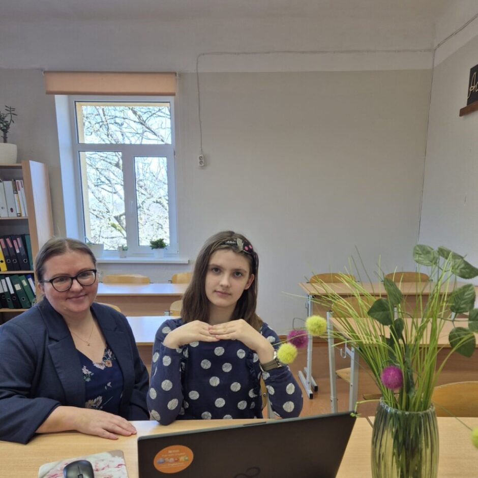 Sieviete un meitene sēž pie koka galda gaišā telpā. Sieviete ar brillēm un tumšu žaketi smaidot skatās kamerā. Meitene, uz galda nolikusi klēpjdatoru, salikusi rokas priekšā un skatās tieši kamerā. Uz galda stāv arī vāze ar zaļiem augiem un krāsainiem pomponu rotājumiem. Fonā redzams logs ar koku zariem un grāmatu plaukts. Meitene ir 4.klases skolniece Sofija Piļipenko, kura piedalās angļu valodas valsts olimpiādē, bet dāma ir viņas skolotāja Linda Liepiņa.