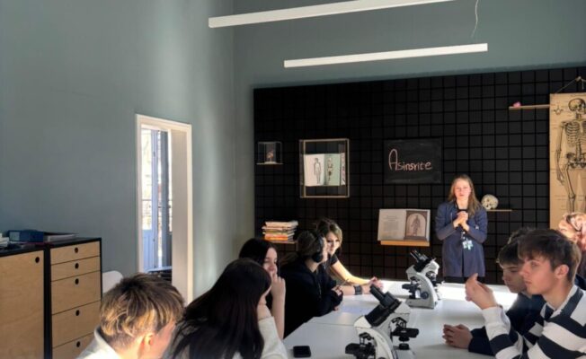 8.klases skolēnu grupa mācību telpā sēž pie gariem galdiem ar mikroskopiem, klausoties pasniedzēju, kas stāv telpas priekšpusē. Uz aizmugurējās sienas redzams cilvēka skeleta attēls un uzraksts 