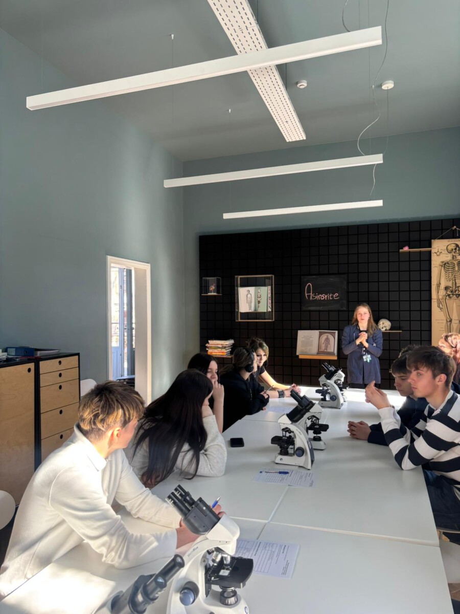 8.klases skolēnu grupa mācību telpā sēž pie gariem galdiem ar mikroskopiem, klausoties pasniedzēju, kas stāv telpas priekšpusē. Uz aizmugurējās sienas redzams cilvēka skeleta attēls un uzraksts 