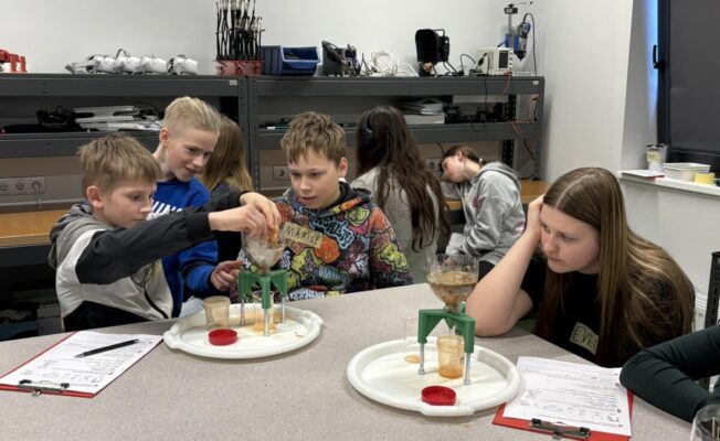 Vairāki 6.,5.klašu skolēni laboratorijas vidē veic eksperimentu. Trīs zēni koncentrējas uz šķidruma liešanu caur piltuvi statīvā. Vienam zēnam ir džemperis ar uzrakstu 