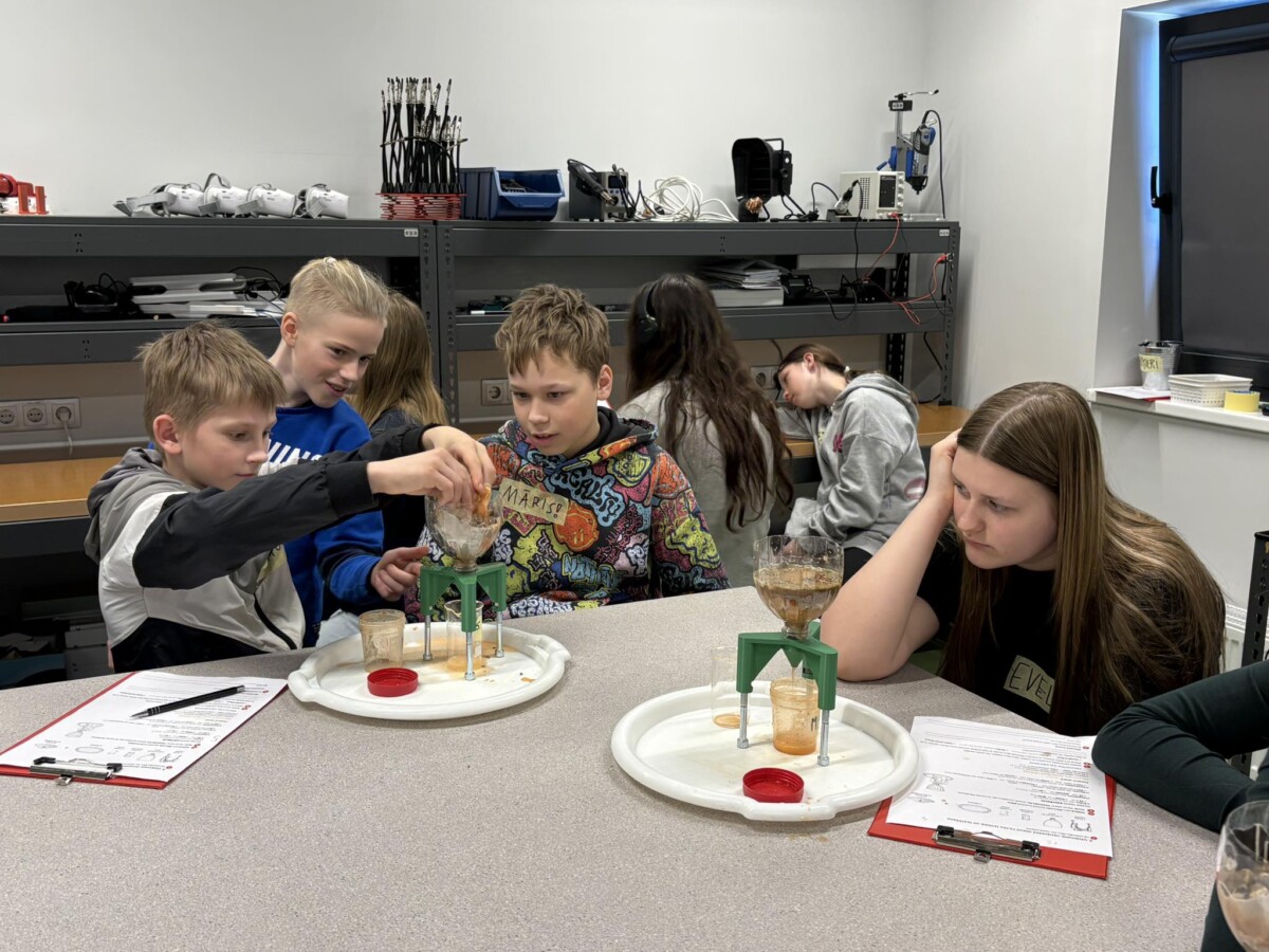 Vairāki 6.,5.klašu skolēni laboratorijas vidē veic eksperimentu. Trīs zēni koncentrējas uz šķidruma liešanu caur piltuvi statīvā. Vienam zēnam ir džemperis ar uzrakstu 