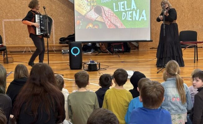 Auditorija ar 1.-9.klašu bērniem sēž uz grīdas, skatoties uz skatuvi. Uz skatuves divi mūziķi uzstājas – viens spēlē akordeonu, otrs vijoli. Aiz viņiem liels ekrāns ar uzrakstu „DARBNĪCA DANČI UN ROTALAS – LIELĀ DIENA”.