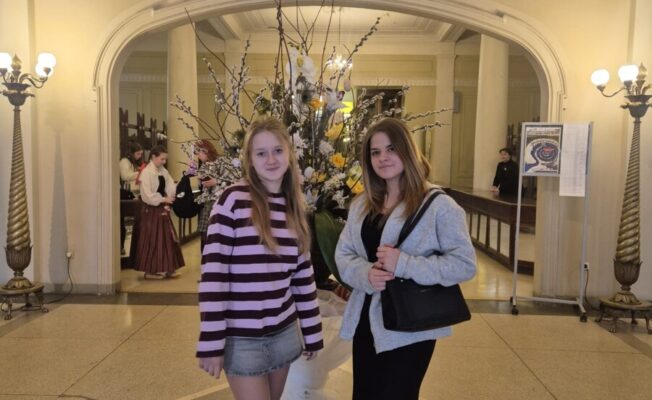 Divas jaunas meitenes stāv greznā zālē, skatoties kamerā. Vienai ir gaiši mati, svītrains violets džemperis un džinsa svārki, otrai – tumši mati, pelēka jaka, melna kleita un melna rokassoma. Starp viņām ir liels ziedu dekors. Fonā redzama arka, virs kuras ir uzraksts: 