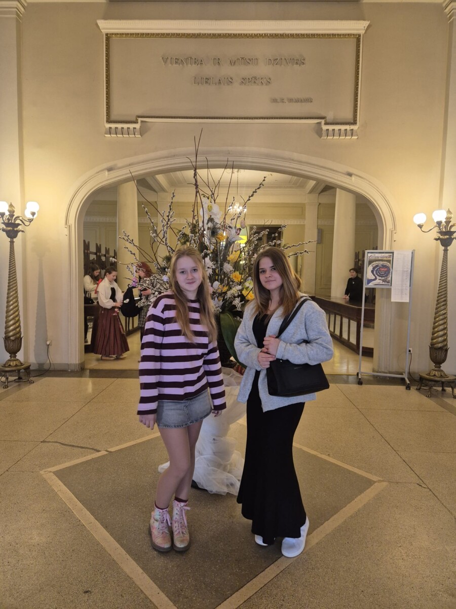 Divas jaunas meitenes stāv greznā zālē, skatoties kamerā. Vienai ir gaiši mati, svītrains violets džemperis un džinsa svārki, otrai – tumši mati, pelēka jaka, melna kleita un melna rokassoma. Starp viņām ir liels ziedu dekors. Fonā redzama arka, virs kuras ir uzraksts: 