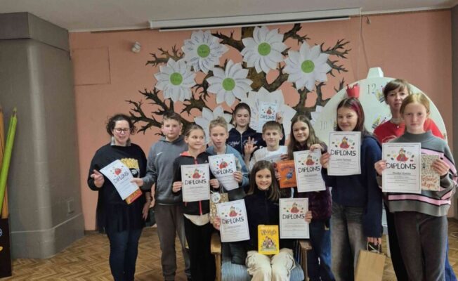 Vairāki 5.klases bērni un skolotājas stāv telpā ar koka grīdu un sienas gleznojumu fonā, turot rokās diplomus ar uzrakstu 