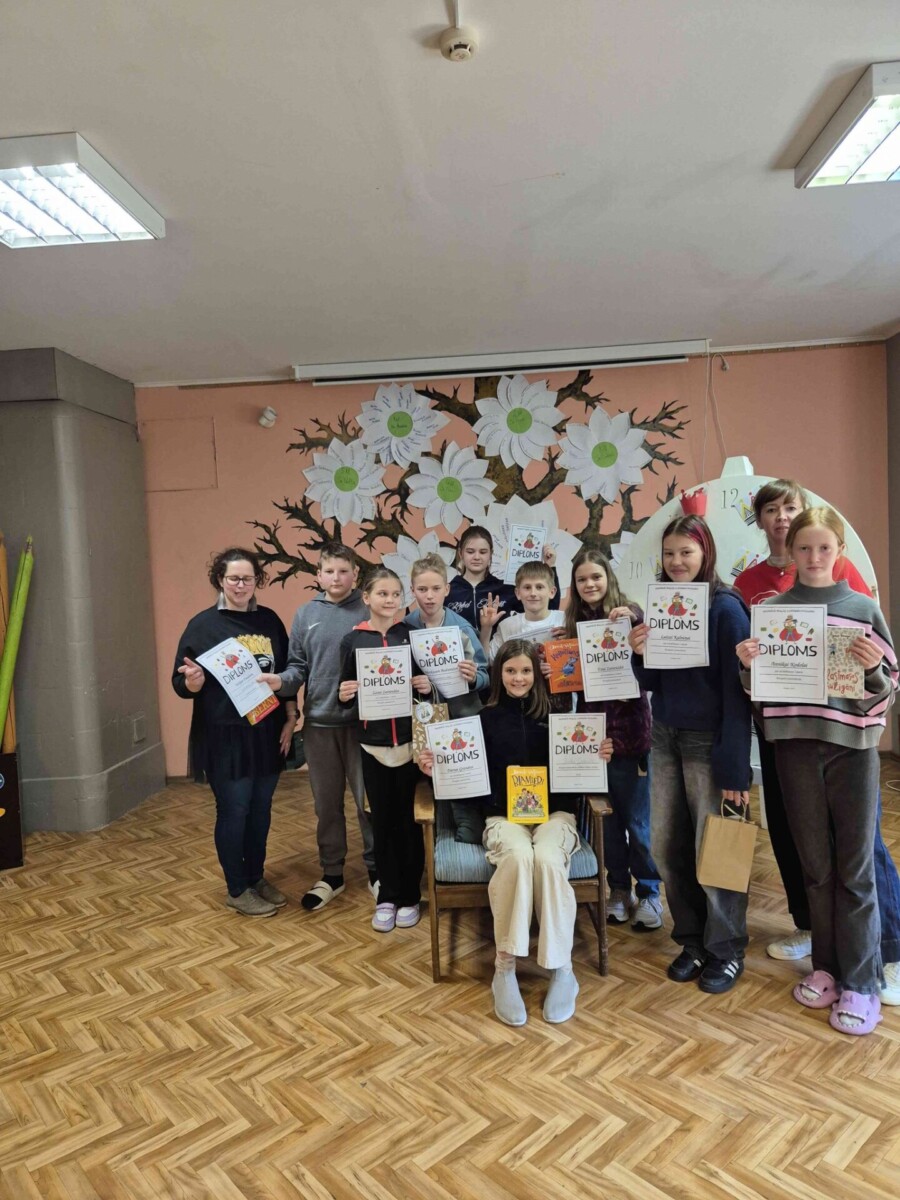 Vairāki 5.klases bērni un skolotājas stāv telpā ar koka grīdu un sienas gleznojumu fonā, turot rokās diplomus ar uzrakstu 