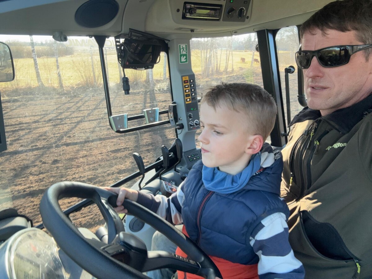 Vīrietis saulesbrillēs un zēns traktorā. Zēns ar rokām uz stūres skatās uz apstrādātu lauku caur logu, bet vīrietis sēž blakus. Zēns 