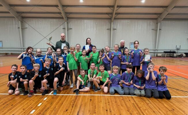 Liela grupa bērnu un četri pieaugušie pozē sporta zālē. Bērni dažādās sporta formās lepni demonstrē medaļas, viens pieaugušais tur kausu, bet cits – diplomu. Visi smaida, skatoties kamerā.