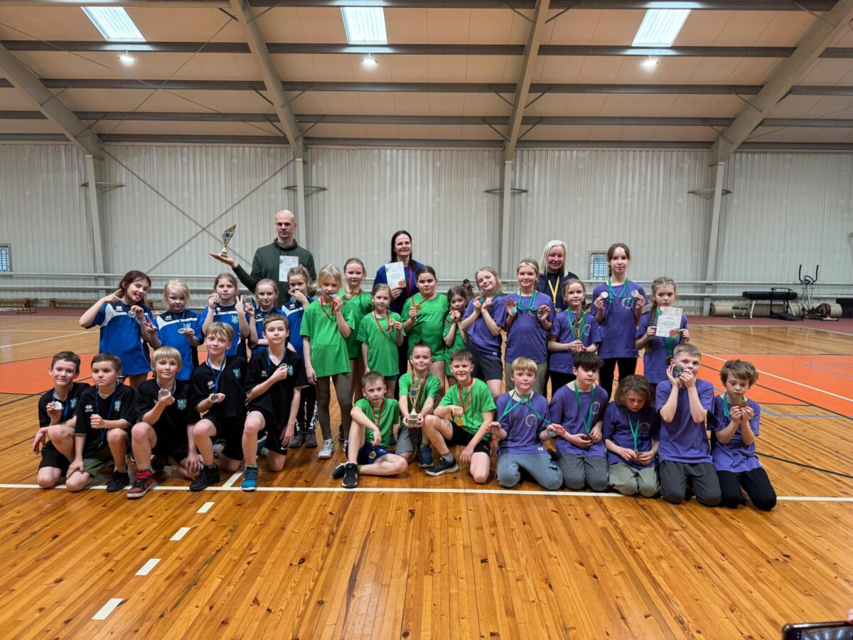 Liela grupa bērnu un četri pieaugušie pozē sporta zālē. Bērni dažādās sporta formās lepni demonstrē medaļas, viens pieaugušais tur kausu, bet cits – diplomu. Visi smaida, skatoties kamerā.