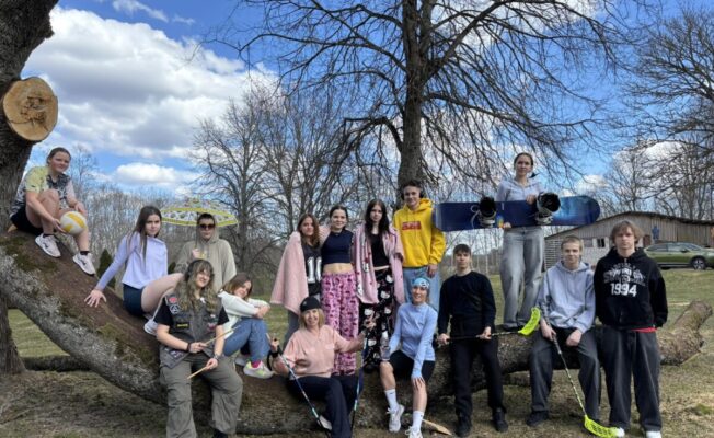 Grupa jauniešu (Birzgales pamatskolas 8.un 9.klašu skolēni) un klašu audzinātājas Baiba Liepiņlauska un Valija Skuja brīvā dabā, daži sēž uz liela nokritušā koka stumbra. Viņi tur dažādu sporta inventāru: volejbola bumbu, snovborda dēli, florbola nūjas un bungu vālītes. Vienam jaunietim mugurā ir džemperis ar uzrakstu 