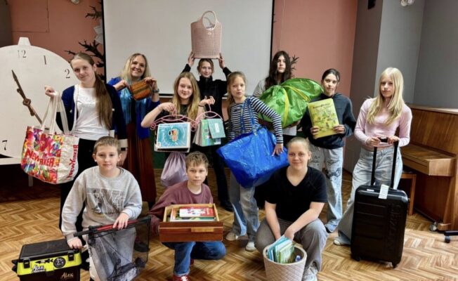 Iekštelpās smaidoša bērnu grupa (6.klase) un viena pieaugušā (Klases audzinātāja Vita Kozlovska), kas pozē ar dažādiem priekšmetiem. Viņi tur krāsainas somas (tostarp zīmola 