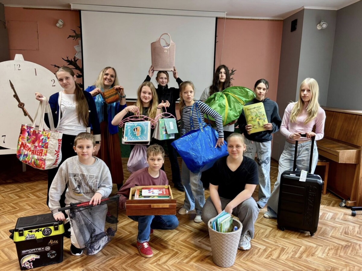 Iekštelpās smaidoša bērnu grupa (6.klase) un viena pieaugušā (Klases audzinātāja Vita Kozlovska), kas pozē ar dažādiem priekšmetiem. Viņi tur krāsainas somas (tostarp zīmola 