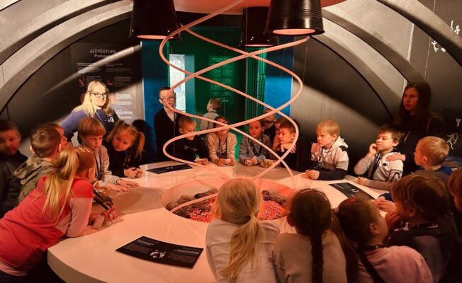 Bērnu grupa un pieaugušie stāv ap apaļu interaktīvu eksponātu zinātnes centrā, kas centrā imitē degošas ogles ar sarkanu spirāli virs tām. Fonā uz sienas redzams stends ar uzrakstu 