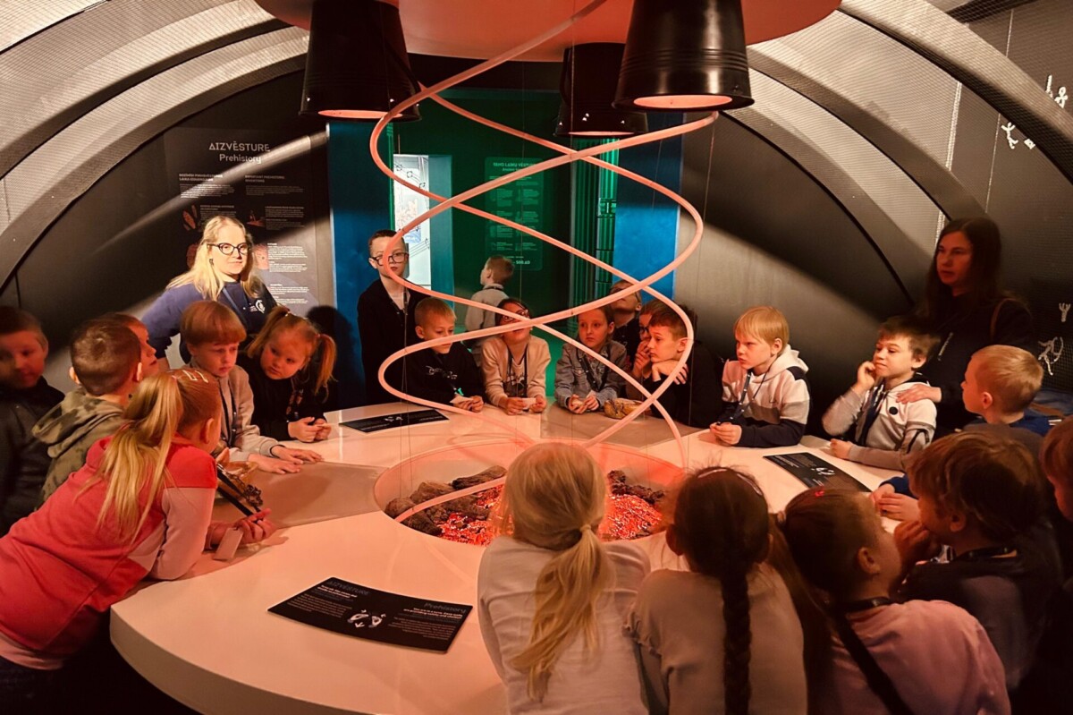 Bērnu grupa un pieaugušie stāv ap apaļu interaktīvu eksponātu zinātnes centrā, kas centrā imitē degošas ogles ar sarkanu spirāli virs tām. Fonā uz sienas redzams stends ar uzrakstu 