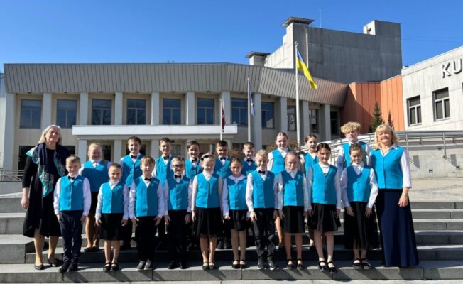 Bērnu 1.-4. klašu koris un skolotāja Sandra Siliņa zilās vestēs un baltos kreklos pozē uz akmens kāpnēm. Fonā ir moderna ēka ar Latvijas un Ukrainas karogiem, uz kuras redzams uzraksts 