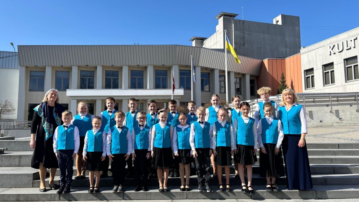 Bērnu 1.-4. klašu koris un skolotāja Sandra Siliņa zilās vestēs un baltos kreklos pozē uz akmens kāpnēm. Fonā ir moderna ēka ar Latvijas un Ukrainas karogiem, uz kuras redzams uzraksts 