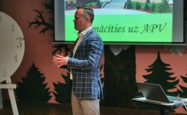 Aizkraukles profesionālās vidusskolas direktors Jānis Siliņš stāv auditorijas priekšā, un prezentē 8.-9.klašu skolēniem šo izglītības iestādi. Aiz viņa uz projektora ekrāna redzama skolas ēka ar logotipu APV un tekstiem 