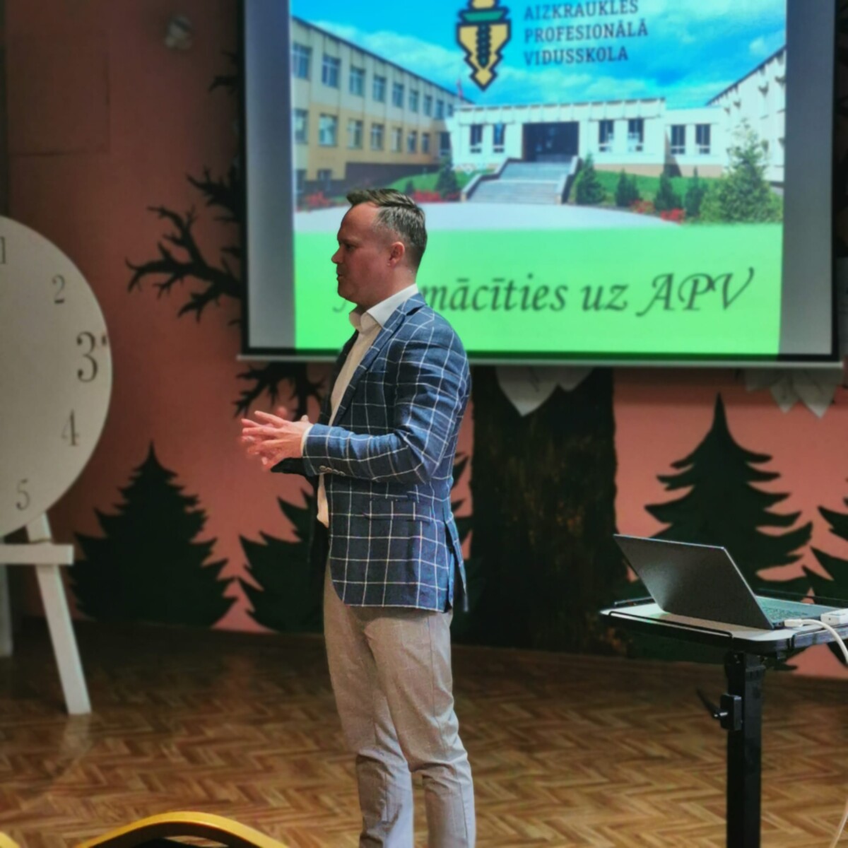 Aizkraukles profesionālās vidusskolas direktors Jānis Siliņš stāv auditorijas priekšā, un prezentē 8.-9.klašu skolēniem šo izglītības iestādi. Aiz viņa uz projektora ekrāna redzama skolas ēka ar logotipu APV un tekstiem 